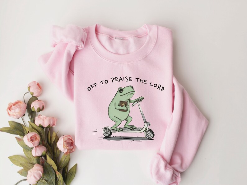 May include: A light pink sweatshirt with the text "OFF TO PRAISE THE LORD" above a cartoon frog riding a scooter while holding a book. The frog is green, and the scooter is white and gray.
