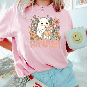 Op de afbeelding: Roze T-shirt met een afbeelding van een spook dat een cocktail drinkt, omringd door bloemen, en het woord "SUMMERWEEN". De persoon houdt een witte mok met een gele smiley vast.