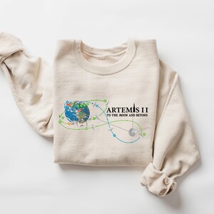 May include: A cream-colored sweatshirt with the text "ARTEMIS II TO THE MOON AND BEYOND" in black. The graphic includes a colorful illustration of Earth, a flight path, and a rocket. The sleeves are rolled up.