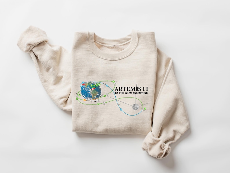 May include: A cream-colored sweatshirt with the text "ARTEMIS II TO THE MOON AND BEYOND" in black. The graphic includes a colorful illustration of Earth, a flight path, and a rocket. The sleeves are rolled up.