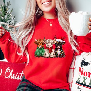 May include: A red sweatshirt displaying three Highland cows in Christmas outfits. One cow is in a green sweater, another in a red scarf, and the third is decorated with fairy lights and a Santa hat. A festive and cosy holiday design.