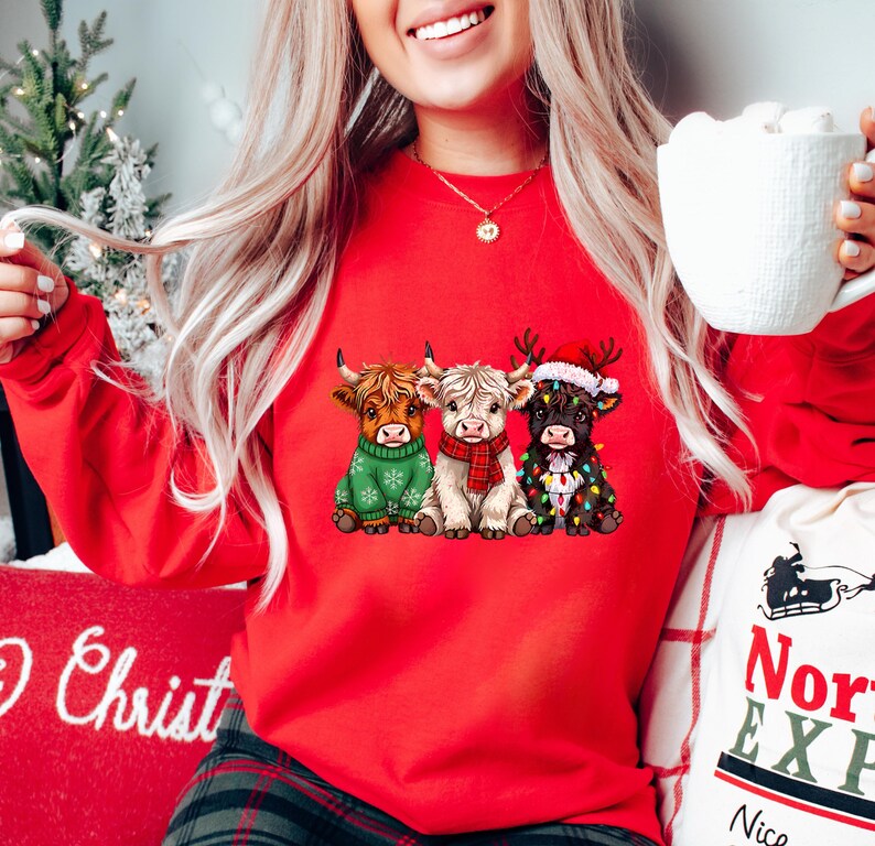 May include: A red sweatshirt displaying three Highland cows in Christmas outfits. One cow is in a green sweater, another in a red scarf, and the third is decorated with fairy lights and a Santa hat. A festive and cosy holiday design.