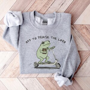 May include: A heather gray sweatshirt with the phrase "OFF TO PRAISE THE LORD" printed above a cartoon frog riding a scooter while holding a book. The frog is green, and the text is black.