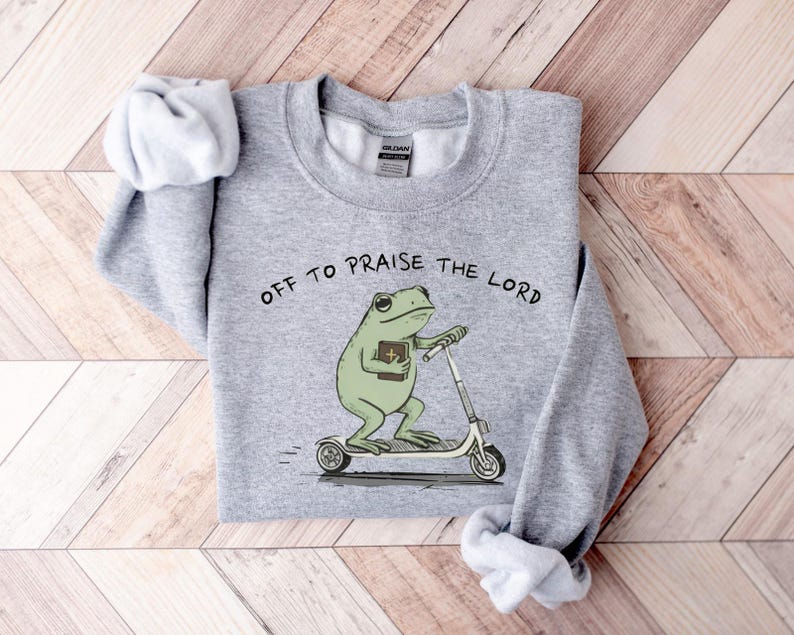 May include: A heather gray sweatshirt with the phrase "OFF TO PRAISE THE LORD" printed above a cartoon frog riding a scooter while holding a book. The frog is green, and the text is black.