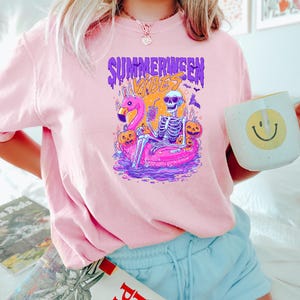 K&ouml;nnte beinhalten: Hellrosa T-Shirt mit einem Grafikdesign, das ein Skelett auf einem Flamingo-Schwimmer zeigt, umgeben von K&uuml;rbissen und Flederm&auml;usen. Der Text "SUMMERWEEN VIBES" ist dar&uuml;ber abgebildet. Eine wei&szlig;e Tasse mit einem gelben Smiley wird gehalten.