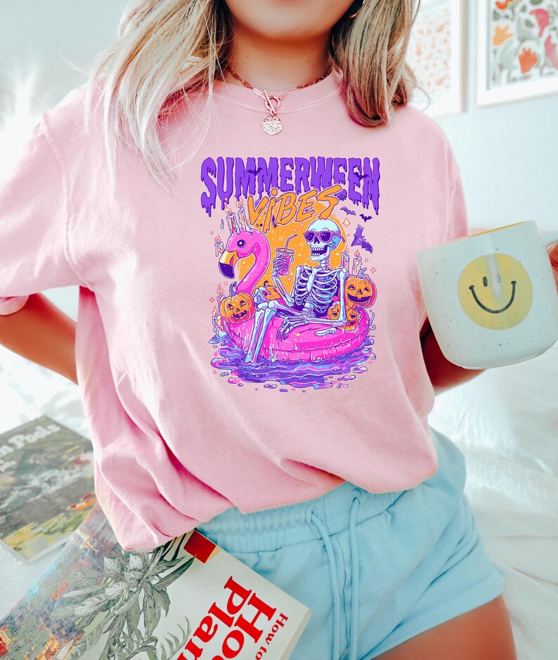 K&ouml;nnte beinhalten: Hellrosa T-Shirt mit einem Grafikdesign, das ein Skelett auf einem Flamingo-Schwimmer zeigt, umgeben von K&uuml;rbissen und Flederm&auml;usen. Der Text "SUMMERWEEN VIBES" ist dar&uuml;ber abgebildet. Eine wei&szlig;e Tasse mit einem gelben Smiley wird gehalten.