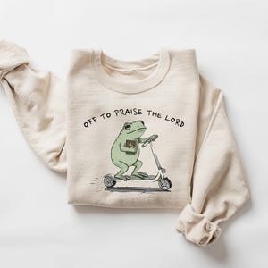 May include: Beige sweatshirt with the text "OFF TO PRAISE THE LORD." The graphic shows a green frog riding a scooter while holding a book. The sweatshirt has long sleeves and a crew neck.