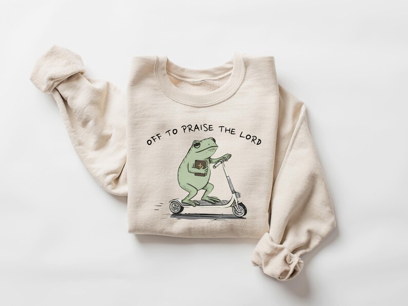 May include: Beige sweatshirt with the text "OFF TO PRAISE THE LORD." The graphic shows a green frog riding a scooter while holding a book. The sweatshirt has long sleeves and a crew neck.