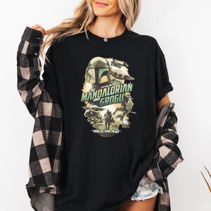 May include: Black t-shirt featuring a graphic from the Star Wars series, "The Mandalorian and Grogu." The design includes images of the Mandalorian helmet, Grogu, and the text "THIS IS THE WAY."