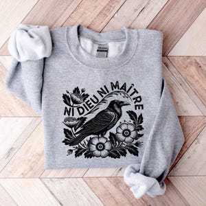 Puede incluir: Sudadera gris jaspeado con un dise&ntilde;o negro de un cuervo rodeado de flores y el texto en franc&eacute;s "Ni Dieu Ni Ma&icirc;tre". La sudadera de cuello redondo y manga larga est&aacute; doblada sobre una superficie de madera.