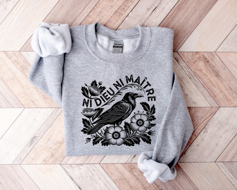 Puede incluir: Sudadera gris jaspeado con un dise&ntilde;o negro de un cuervo rodeado de flores y el texto en franc&eacute;s "Ni Dieu Ni Ma&icirc;tre". La sudadera de cuello redondo y manga larga est&aacute; doblada sobre una superficie de madera.
