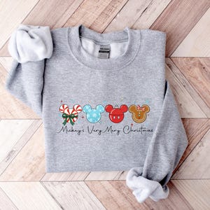 May include: A light gray sweatshirt featuring a festive design. The design includes candy canes, Mickey Mouse heads, and gingerbread men, all in Christmas colors. The text "Mickey's Very Merry Christmas" is written below the design.
