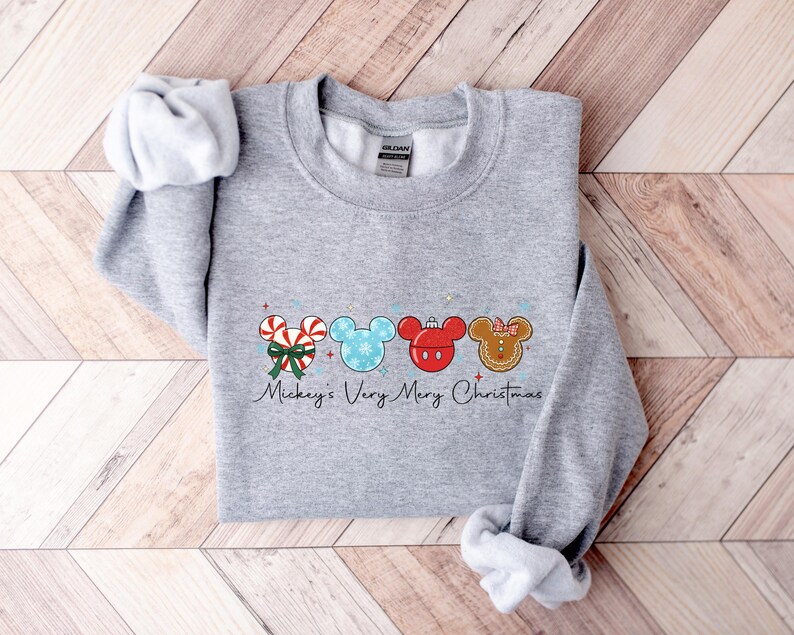 May include: A light gray sweatshirt featuring a festive design. The design includes candy canes, Mickey Mouse heads, and gingerbread men, all in Christmas colors. The text "Mickey's Very Merry Christmas" is written below the design.