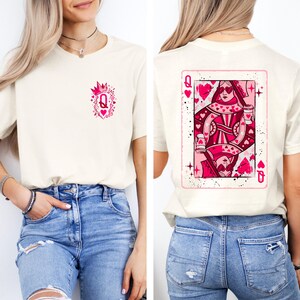 May include: Cream-colored t-shirt featuring a pink Queen of Hearts playing card design on the back and a smaller matching design on the front. The card design includes hearts and the letter "Q". The shirt is made of a soft material.