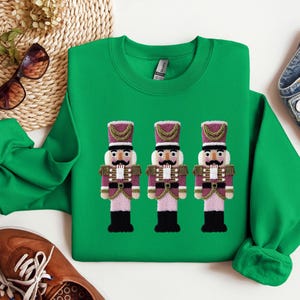 May include: A green sweatshirt featuring three embroidered nutcracker figures. The nutcrackers have purple and gold accents, black boots, and white mustaches. The sweatshirt is a crew neck style.