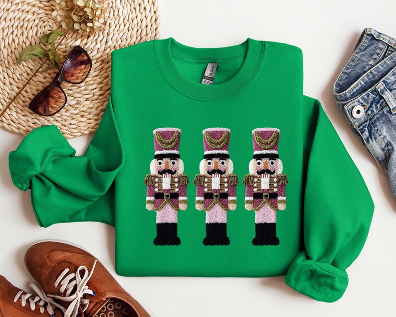 May include: A green sweatshirt featuring three embroidered nutcracker figures. The nutcrackers have purple and gold accents, black boots, and white mustaches. The sweatshirt is a crew neck style.
