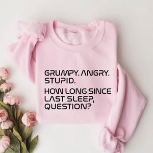 May include: A light pink sweatshirt with the text "GRUMPY. ANGRY. STUPID. HOW LONG SINCE LAST SLEEP, QUESTION?" in black. The sweatshirt is folded, with a bouquet of pink flowers in the lower left corner.