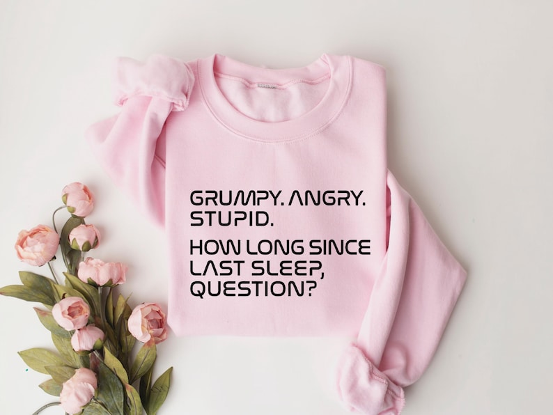 May include: A light pink sweatshirt with the text "GRUMPY. ANGRY. STUPID. HOW LONG SINCE LAST SLEEP, QUESTION?" in black. The sweatshirt is folded, with a bouquet of pink flowers in the lower left corner.