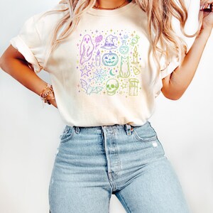 May include: Cream-colored t-shirt featuring a colorful Halloween-themed graphic. The design includes a ghost, witch hat, pumpkin, cat, skull, and other spooky elements. The shirt is paired with light blue denim jeans.
