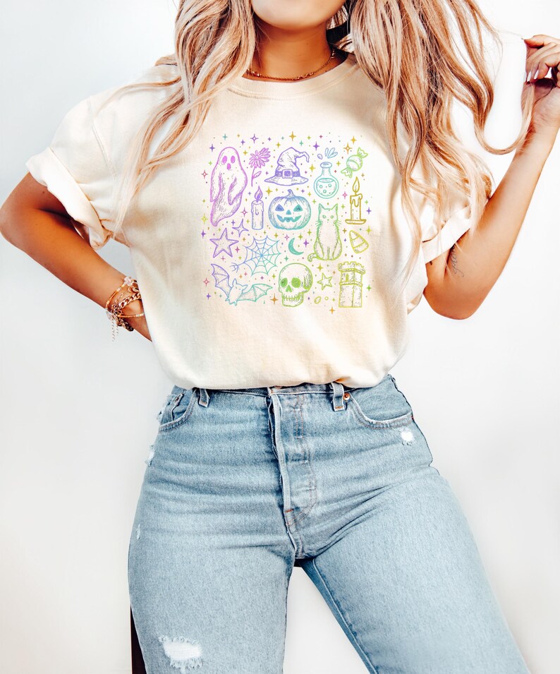 May include: Cream-colored t-shirt featuring a colorful Halloween-themed graphic. The design includes a ghost, witch hat, pumpkin, cat, skull, and other spooky elements. The shirt is paired with light blue denim jeans.