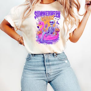K&ouml;nnte beinhalten: Cremefarbenes T-Shirt mit dem Text "Summerween Vibes" in Lila und Orange. Die Grafik zeigt ein Skelett, das sich auf einem rosa Flamingo-Schwimmer entspannt, umgeben von K&uuml;rbissen und Flederm&auml;usen. Das Skelett h&auml;lt ein Getr&auml;nk.