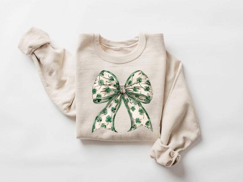 May include: A cream-colored sweatshirt featuring a large bow graphic. The bow is detailed with green shamrocks on a white background, with green trim. The sweatshirt has long sleeves and a crew neck.