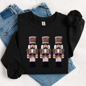 May include: Black sweatshirt featuring three embroidered nutcracker figures. The nutcrackers have pink and burgundy uniforms with gold accents, black boots, and white faces. The sweatshirt is displayed on top of blue jeans.