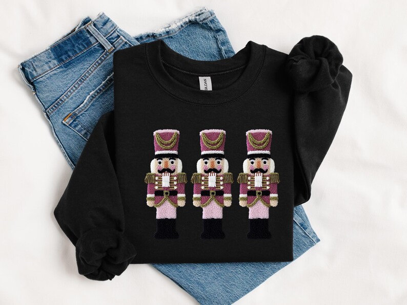 May include: Black sweatshirt featuring three embroidered nutcracker figures. The nutcrackers have pink and burgundy uniforms with gold accents, black boots, and white faces. The sweatshirt is displayed on top of blue jeans.