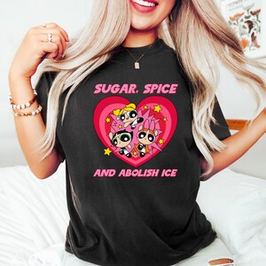 May include: Black t-shirt featuring a graphic of three cartoon characters inside a pink heart with the text "SUGAR, SPICE" above and "AND ABOLISH ICE" below. The characters have different hair colors and expressions.