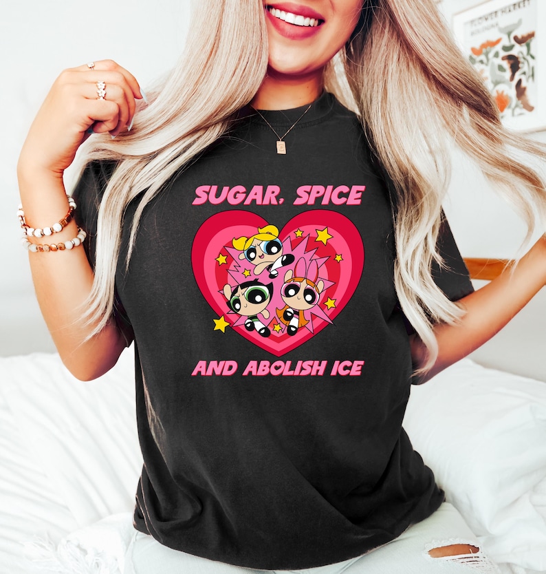 May include: Black t-shirt featuring a graphic of three cartoon characters inside a pink heart with the text "SUGAR, SPICE" above and "AND ABOLISH ICE" below. The characters have different hair colors and expressions.
