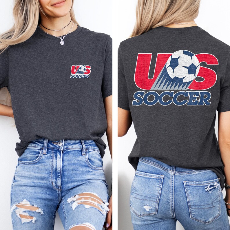 Puede incluir: Una camiseta de manga corta en color gris carb&oacute;n con un peque&ntilde;o logotipo de US Soccer en el pecho izquierdo y un gr&aacute;fico grande en la espalda. El dise&ntilde;o trasero muestra el texto US en rojo con un bal&oacute;n de f&uacute;tbol y la palabra SOCCER en letras azules.