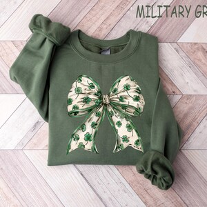 May include: Military green sweatshirt featuring a large bow with shamrock and ribbon design. The bow has a cream background with green shamrocks and green ribbon trim. The sweatshirt is on a light wood grain background.