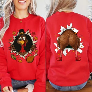May include: Red sweatshirt with a Thanksgiving turkey design. The front features a turkey wearing a pilgrim hat bursting through a torn hole, surrounded by fall leaves and corn. The back shows the turkey's rear and legs.