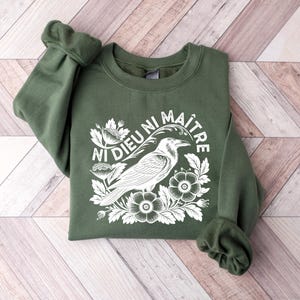 Puede incluir: Una sudadera verde oliva con el texto "Ni Dieu Ni Ma&icirc;tre" y un gr&aacute;fico blanco de un cuervo, flores y follaje. La sudadera est&aacute; doblada sobre una superficie de madera marr&oacute;n claro y blanca.