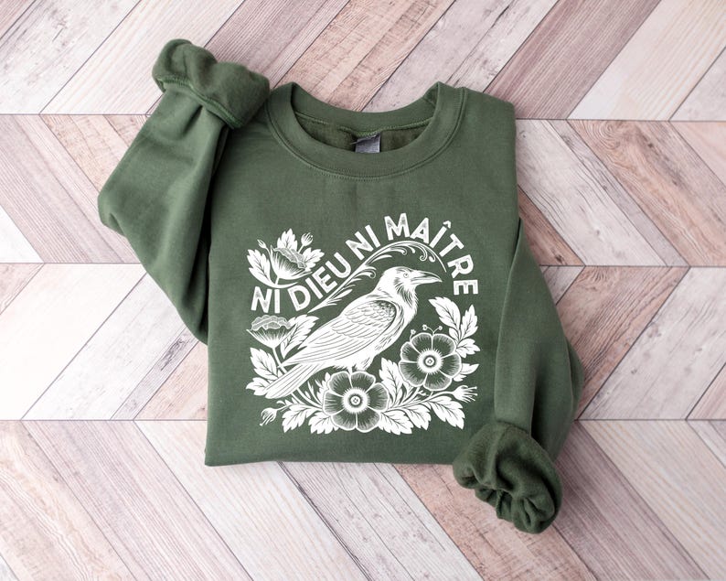 Puede incluir: Una sudadera verde oliva con el texto "Ni Dieu Ni Ma&icirc;tre" y un gr&aacute;fico blanco de un cuervo, flores y follaje. La sudadera est&aacute; doblada sobre una superficie de madera marr&oacute;n claro y blanca.