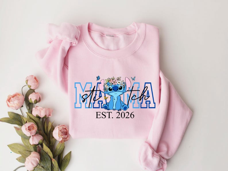 May include: A light pink sweatshirt with the words "Mama Stitch" in blue and white lettering, featuring a cartoon character with a floral crown. The text "EST. 2026" is below the character.