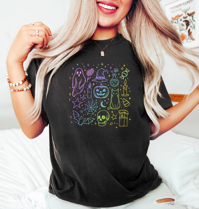 May include: Black t-shirt featuring a colorful Halloween-themed graphic. The design includes a ghost, witch hat, pumpkin, cat, skull, and other spooky elements. The graphic is in a square shape and has a rainbow gradient.
