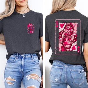 May include: Dark gray t-shirt featuring a pink graphic of a queen of hearts playing card on the back. The front has a small pink design with a crown and the letter "Q". The shirt is paired with blue jeans.