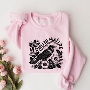 Puede incluir: Sudadera rosa claro con un dise&ntilde;o de cuervo negro rodeado de flores y la frase francesa "Ni Dieu Ni Ma&icirc;tre". El dise&ntilde;o est&aacute; centrado en la parte delantera de la sudadera de cuello redondo.