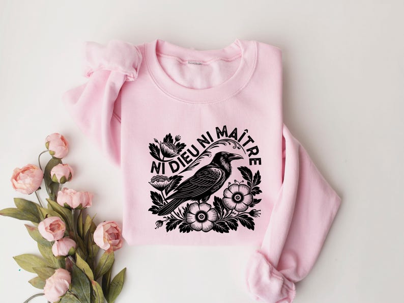 Puede incluir: Sudadera rosa claro con un dise&ntilde;o de cuervo negro rodeado de flores y la frase francesa "Ni Dieu Ni Ma&icirc;tre". El dise&ntilde;o est&aacute; centrado en la parte delantera de la sudadera de cuello redondo.