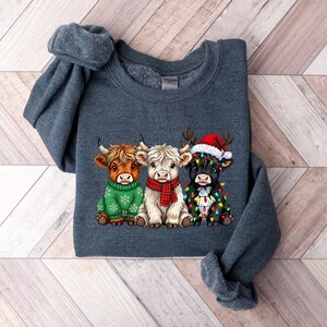 May include: A dark grey sweatshirt with a graphic of three Highland cows. One cow is wearing a green sweater, another a red scarf, and the third a Santa hat and Christmas lights. The cows are cartoon-style.