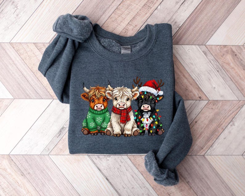 May include: A dark grey sweatshirt with a graphic of three Highland cows. One cow is wearing a green sweater, another a red scarf, and the third a Santa hat and Christmas lights. The cows are cartoon-style.