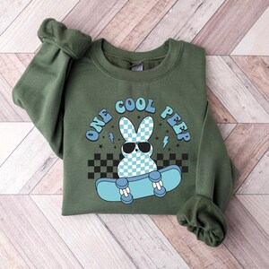 May include: Green sweatshirt featuring the text "ONE COOL PEEP" and a cartoon bunny wearing sunglasses on a skateboard. The bunny has a blue and white checkered pattern. The skateboard is light blue with black and white checkered accents.
