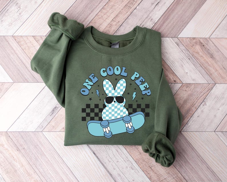 May include: Green sweatshirt featuring the text "ONE COOL PEEP" and a cartoon bunny wearing sunglasses on a skateboard. The bunny has a blue and white checkered pattern. The skateboard is light blue with black and white checkered accents.