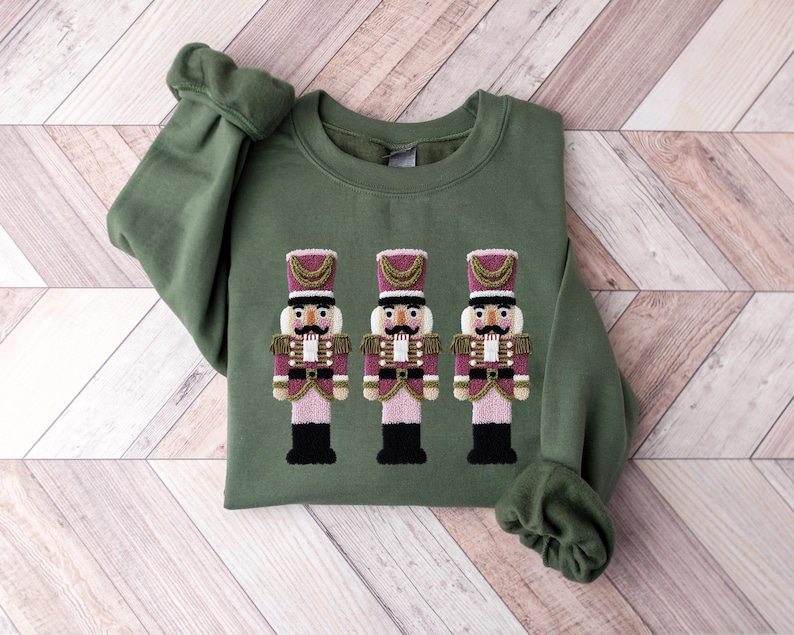 May include: An olive green sweatshirt featuring an embroidered design of three toy nutcracker soldiers. The nutcrackers have pink and purple uniforms with gold accents and black boots. The sweatshirt has long sleeves and a crew neck.