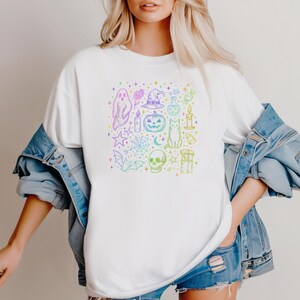 May include: White t-shirt featuring a colorful Halloween-themed graphic. The design includes a ghost, witch hat, pumpkin, cat, skull, and other spooky elements. The graphic is in a rainbow gradient color scheme.