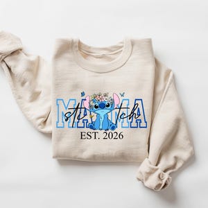 May include: Beige sweatshirt featuring a blue cartoon character with a floral crown and the text "Stitch Mama EST. 2026" in blue. The sweatshirt has rolled-up sleeves.