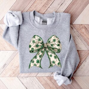 May include: A light gray sweatshirt featuring a large bow design. The bow has a cream-colored background with green shamrocks and green trim. The sweatshirt is made of a soft material and is perfect for St. Patrick's Day.
