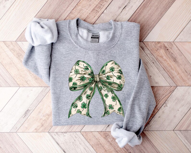 May include: A light gray sweatshirt featuring a large bow design. The bow has a cream-colored background with green shamrocks and green trim. The sweatshirt is made of a soft material and is perfect for St. Patrick's Day.