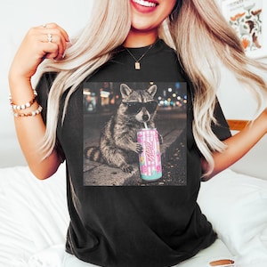 May include: Black t-shirt featuring a graphic of a raccoon wearing sunglasses and holding a pink can with the text "Fluff". The raccoon is sitting on a curb with a city background. The can has a floral and striped design.